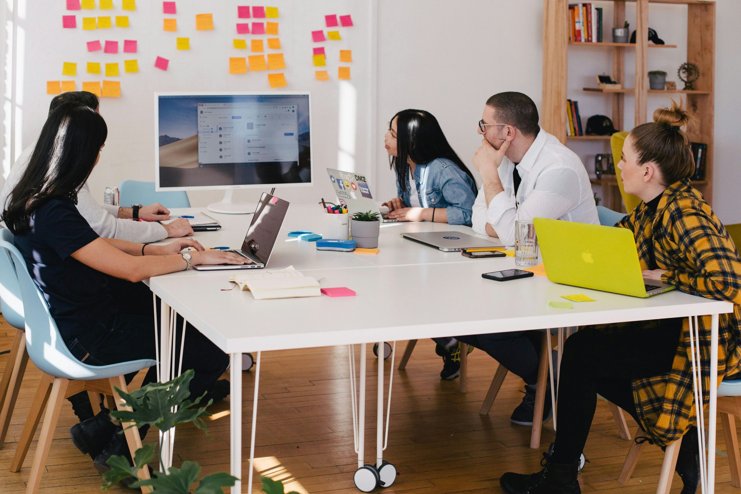 Group gathered around a desk having a meeting in an office. Wall in background covered with bright multi coloured post-it notes.